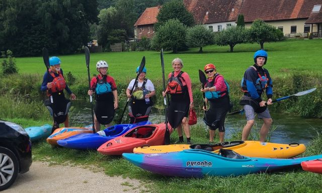 6 Personen in Kanuoutfit stehen hinter bunten Kajaks am Brenzufer im Eselsburger Tal